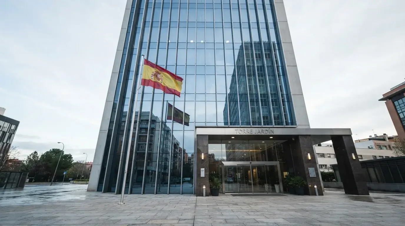 Fachada de un edificio de oficinas con bandera española ondeando junto a la entrada principal de cristal.